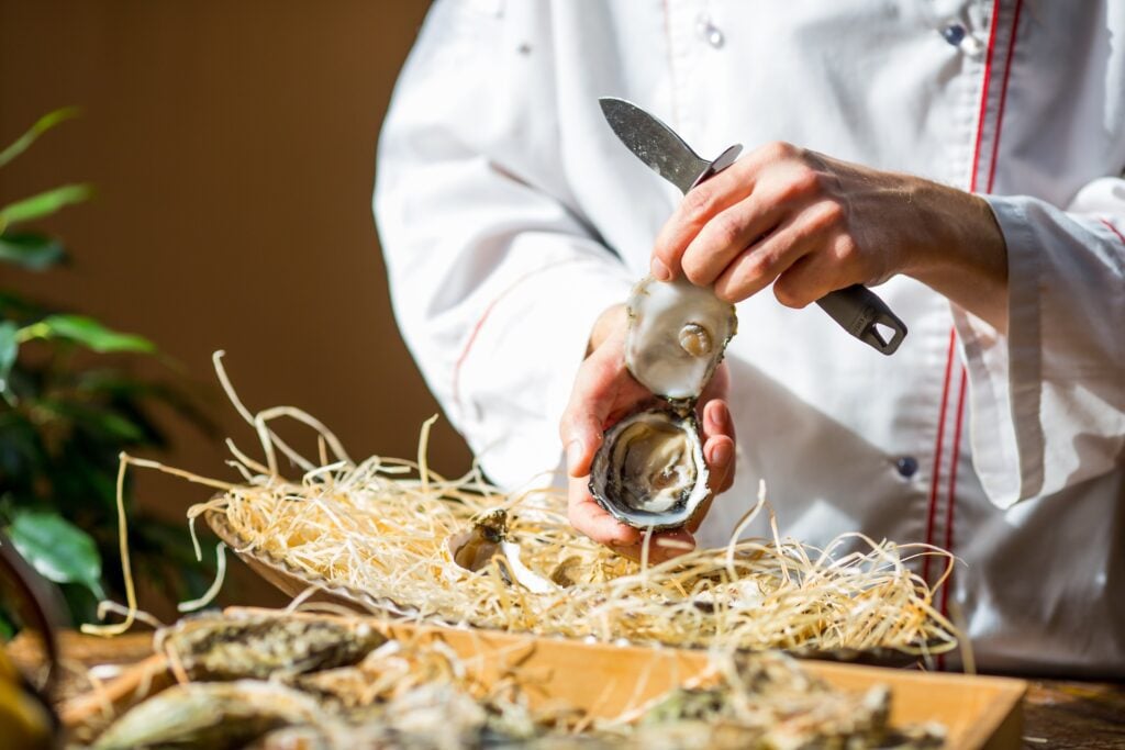 Chef opening a fresh oyster with a knife in a professional kitchen.