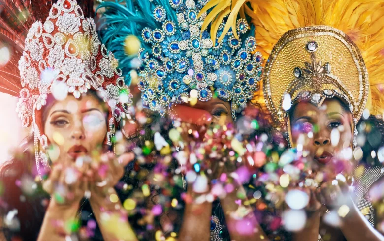 Three women in vibrant feathered Mardi Gras costumes blowing confetti